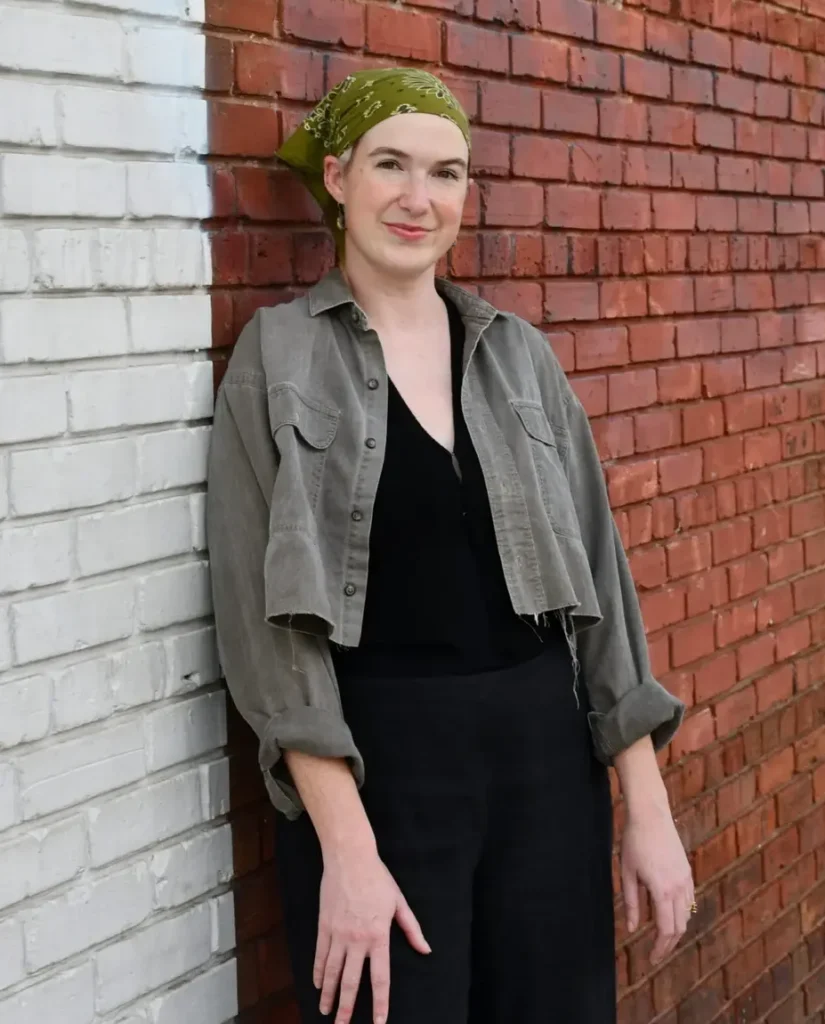 A woman in a green headscarf and jacket stands against a white and red brick background.
