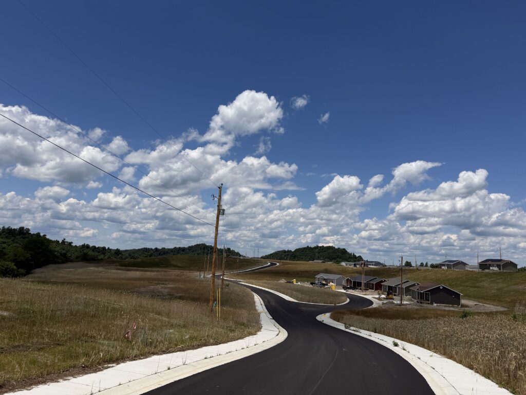 Curving freshly paved road through a grassy, open area with houses along the right and a utility pole with power lines against a bright blue sky with clouds.