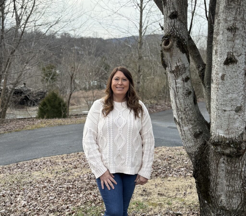Smiling woman standing beside a large tree outdoors, wearing a cream sweater and blue jeans, with fallen leaves on the ground.