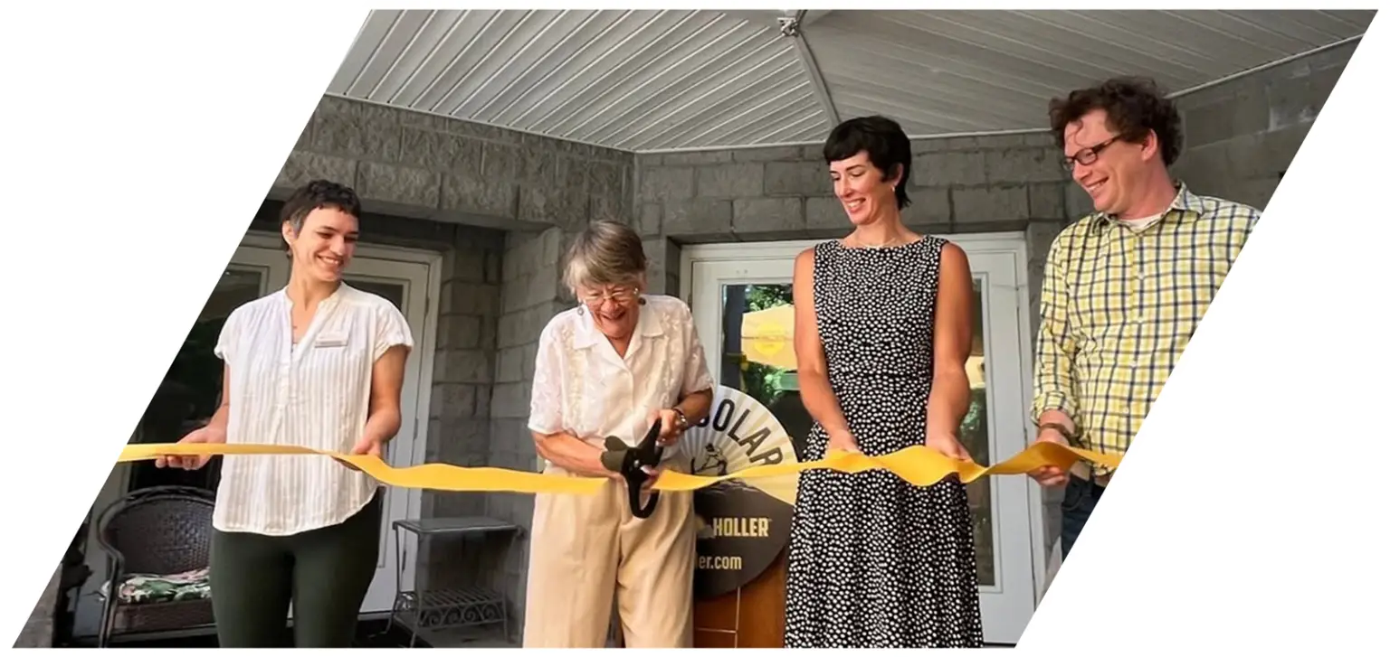 Four persons holding a yellow ribbon during a ribbon cutting ceremony in front a building entrance.